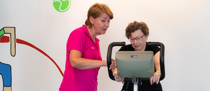 Mevrouw krijgt uitleg van fysiotherapeut over fitnessapparaat